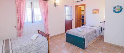 Une chambre avec deux lits, une fenêtre avec des rideaux, un climatiseur fixé au mur et une porte donnant sur une autre pièce.