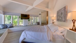 A bedroom with a large bed, a TV mounted on the wall, and a window with a view of greenery.