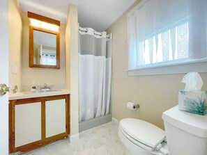 A bathroom with a wooden vanity, a toilet, a shower with a white curtain, and a window with patterned curtains. - Seascapes (Wells)