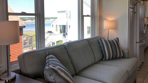 A living room with a sofa, two pillows, a lamp, and a view of a house and water through large windows.