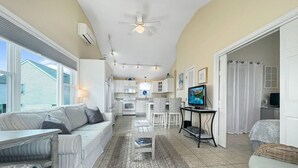 A spacious living area with a sofa, a coffee table, and a television. There is a kitchen area with a microwave and a refrigerator. The room has large windows and a ceiling fan.