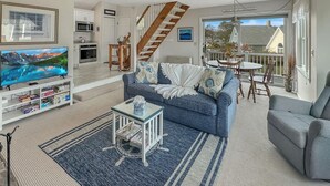 Un salón amplio con un sofá azul, una mesa de centro y un televisor sobre un mueble bajo. Una escalera conduce a un nivel superior.