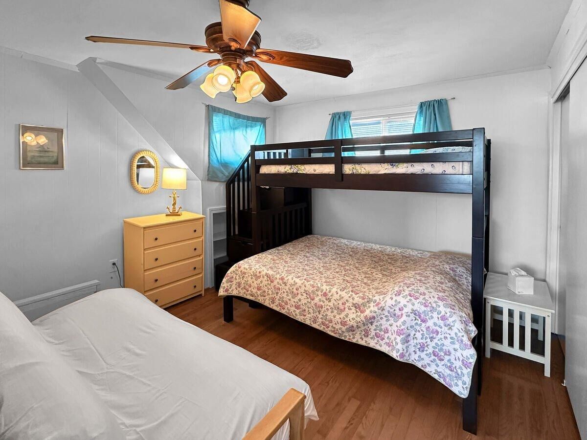 A bedroom with a bunk bed, a single bed, a wooden dresser, a ceiling fan, and a window with curtains.