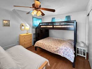 A bedroom with a bunk bed, a single bed, a wooden dresser, a ceiling fan, and a window with curtains. - Sweet Serenity (York Beach)