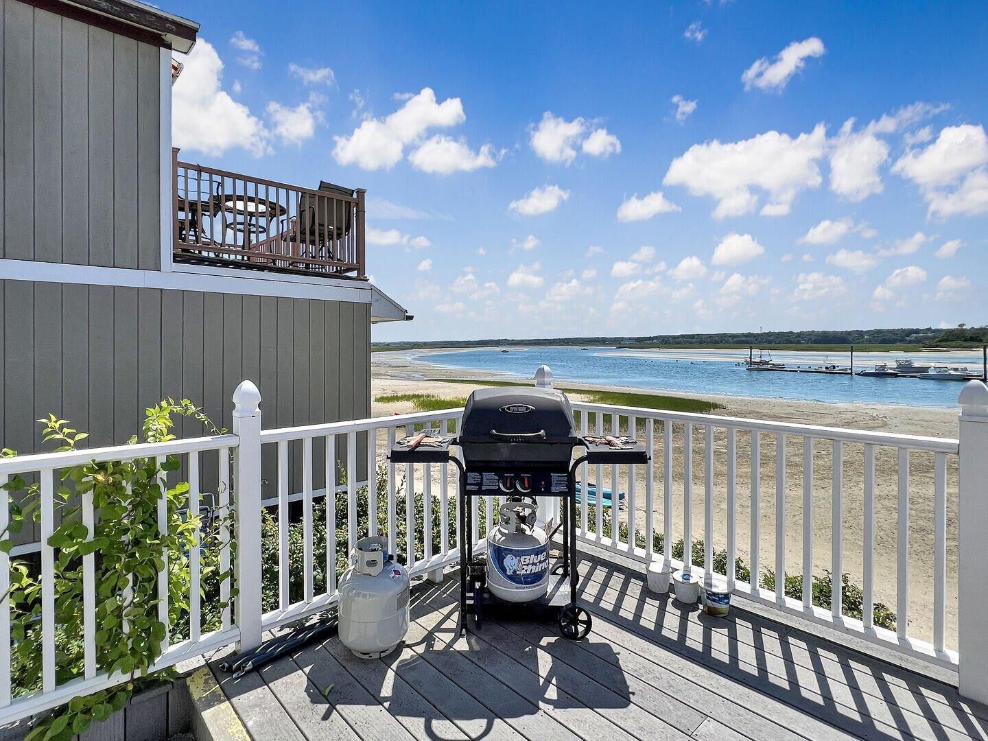 A deck with a grill, propane tank, and outdoor furniture overlooking a beach.