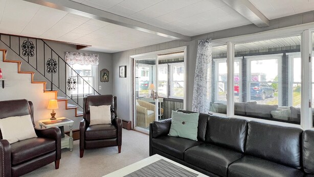 A living room with a sofa, armchair, and a coffee table. There is a staircase with decorative railings and a view of the outside through large windows. - Sweet Dreams (Wells)