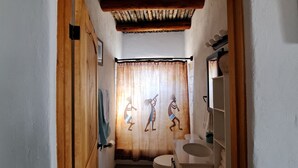 A narrow hallway leading to a bathroom with a wooden door, a towel hanging on the wall, and a shower curtain with tribal patterns. - Cozy Spanish Casita (El Prado)