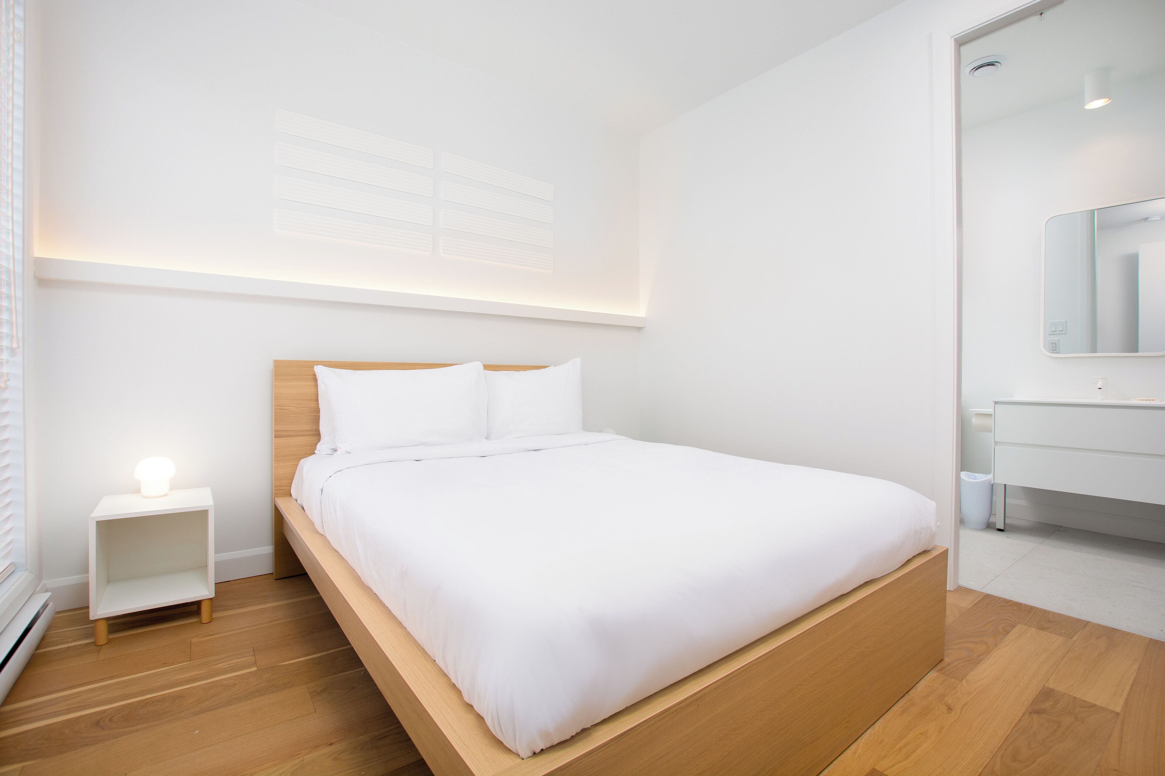 Ein modernes Schlafzimmer mit einem hölzernen Bett, weißer Bettwäsche, einem Nachttisch mit Lampe und einer weißen Kommode.