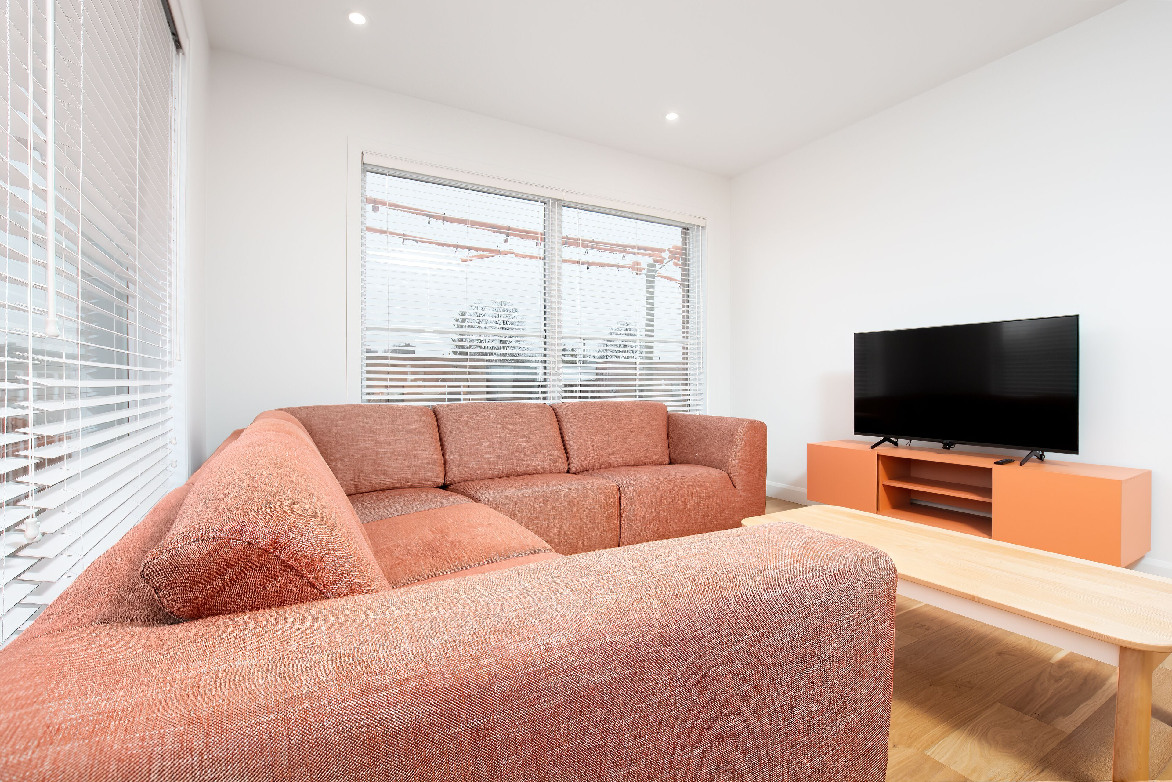 Ein modernes Wohnzimmer mit einem Ecksofa, einem Flachbildfernseher auf einem Holzboden und einem großen Fenster mit Jalousien.