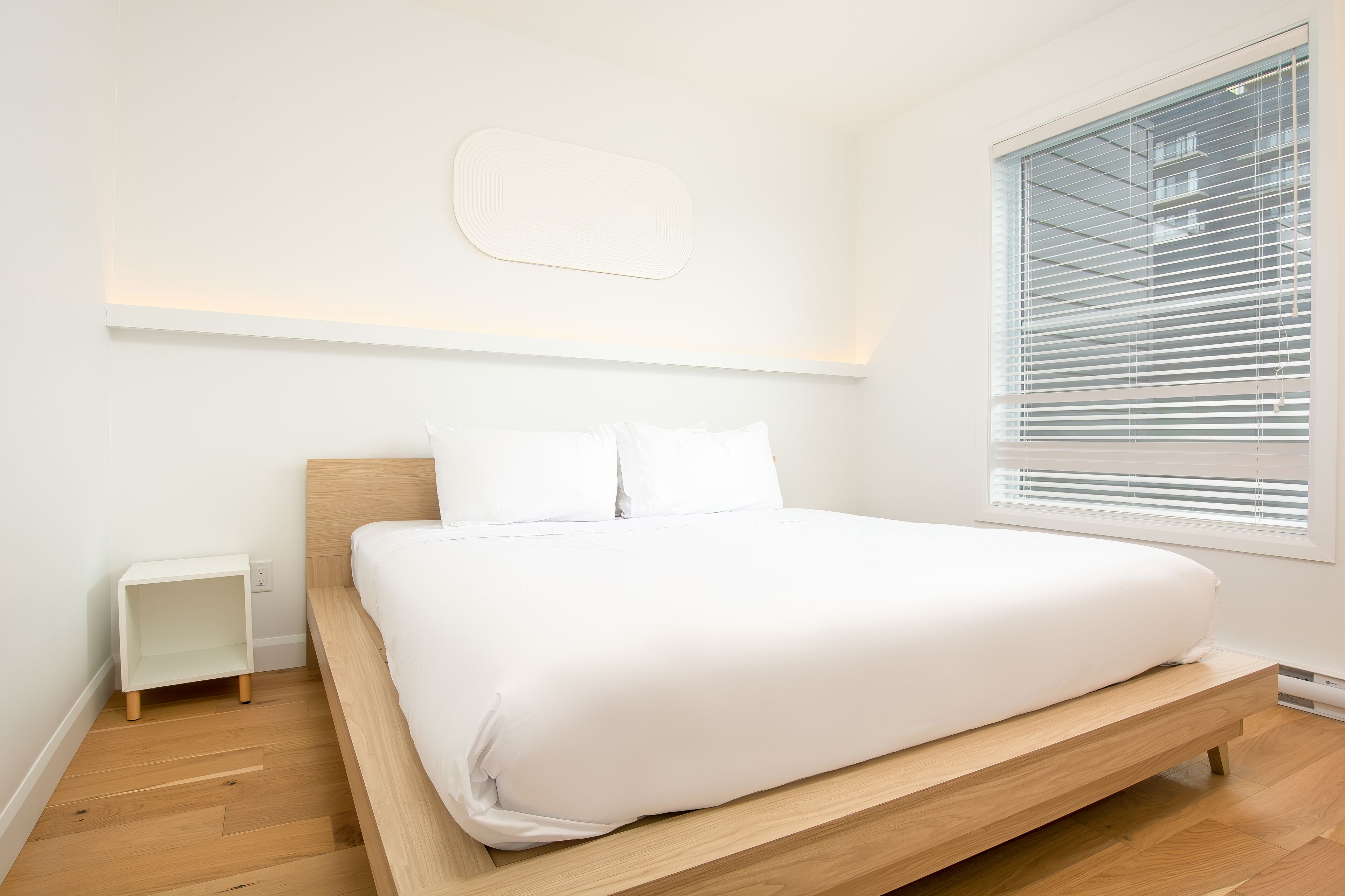 A minimalist bedroom with a wooden bed, white bedding, a small bedside table, and a large window with blinds.