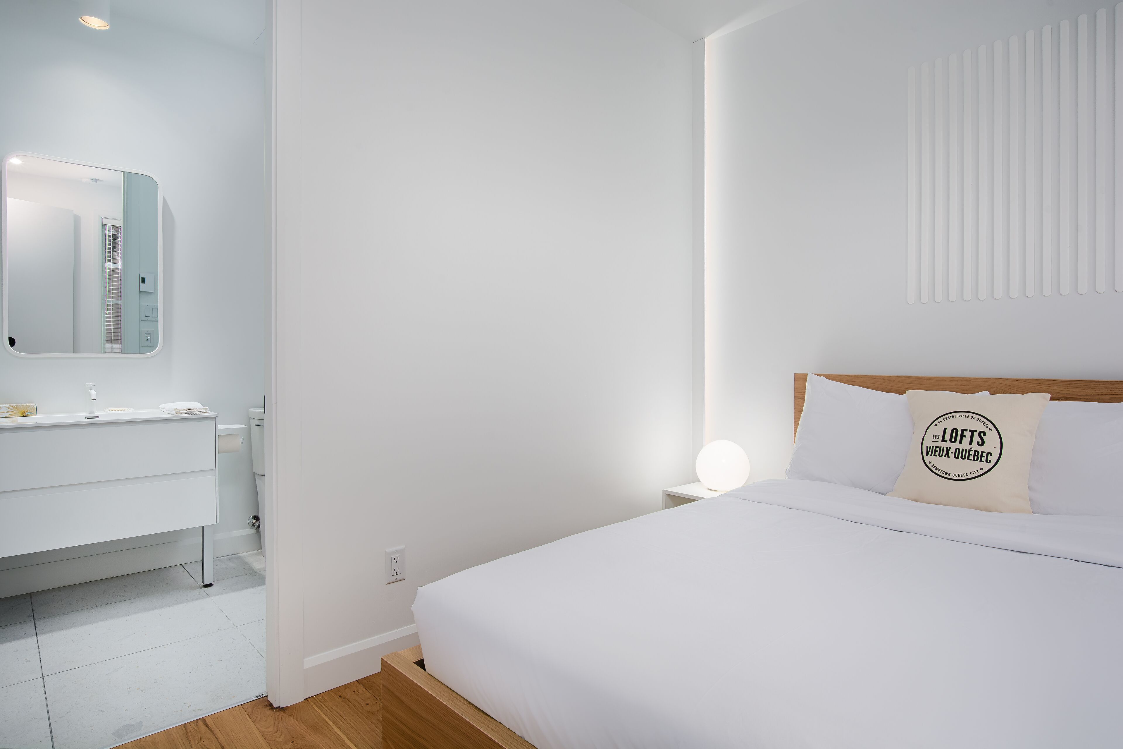 Et moderne hotelværelse med en hvid seng, et natbord med lampe, et badeværelse med spejl og håndvask samt en radiator monteret på væggen.
