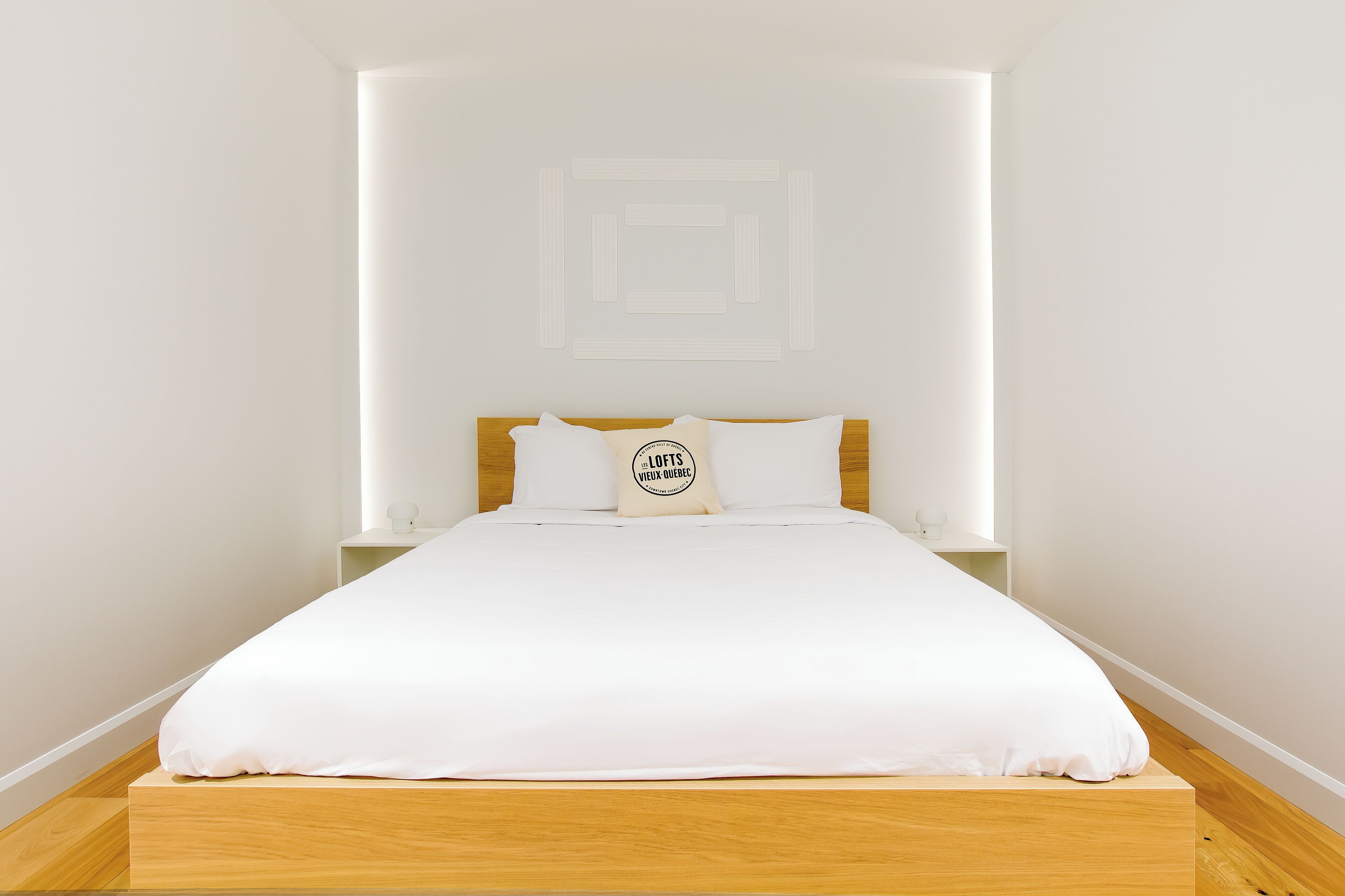 A neatly made bed with white bedding and a pillow with a logo, a wooden headboard, and a white wall with a geometric design.