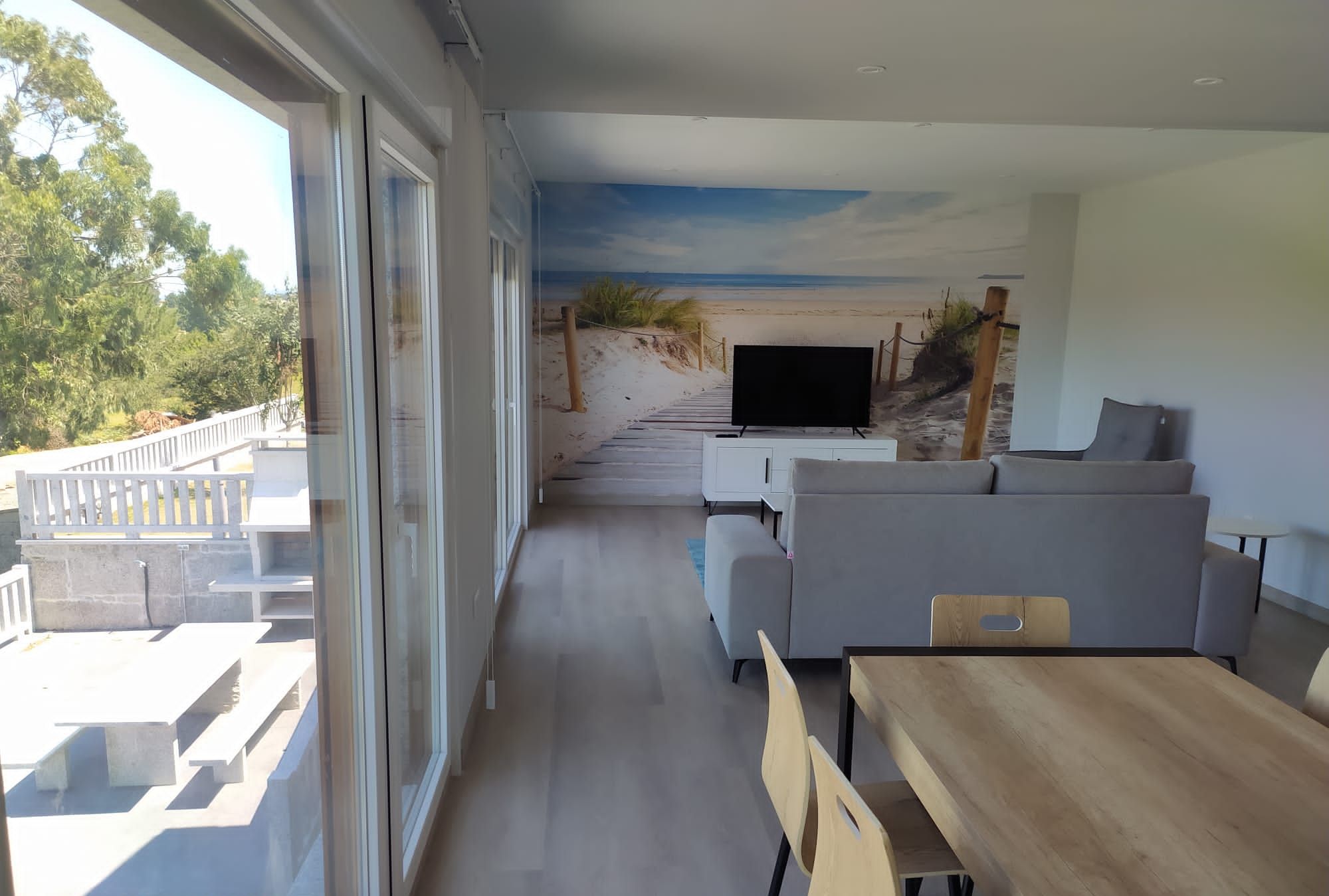 Een moderne woonkamer met een grote televisie, een grijze bank, een houten eettafel en stoelen.