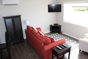 A living room with a red sofa, a TV mounted on the wall, a small table with remotes, and a window with a view of greenery.