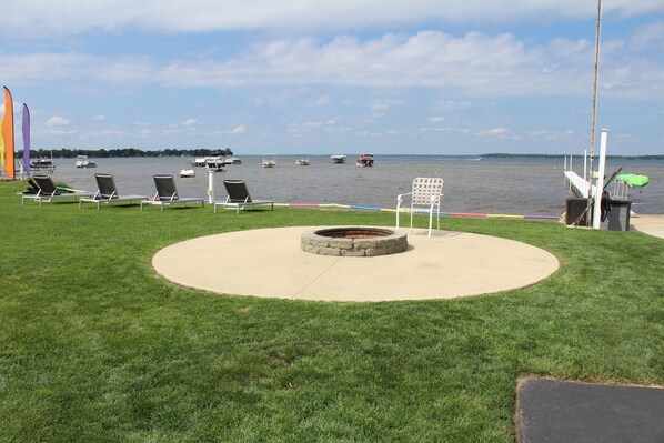 A lakeside patio with lounge chairs, a fire pit, and a view of boats on the water.