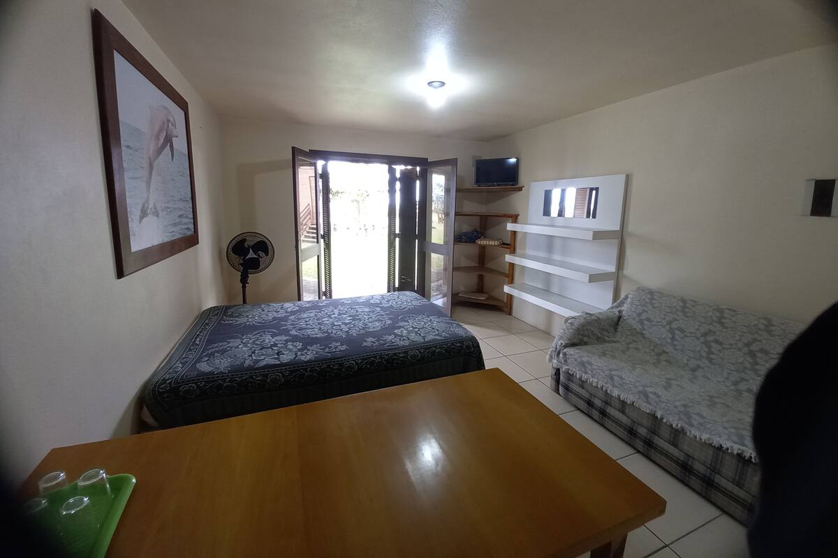 Ein Zimmer mit einem Bett, einem Holztisch, einer Couch, einem Fernseher auf einem Regal und einem Ventilator.