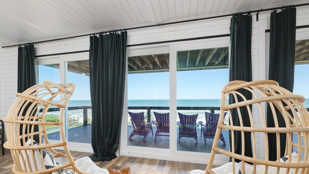 A room with wicker chairs, a wooden table, and a large window overlooking the beach.
