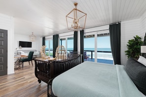A bedroom with a large bed, a desk with a chair, a television, and a view of the ocean. - Oceanfront 8/4 | Industrial Chic | Large Groups (St. Augustine)