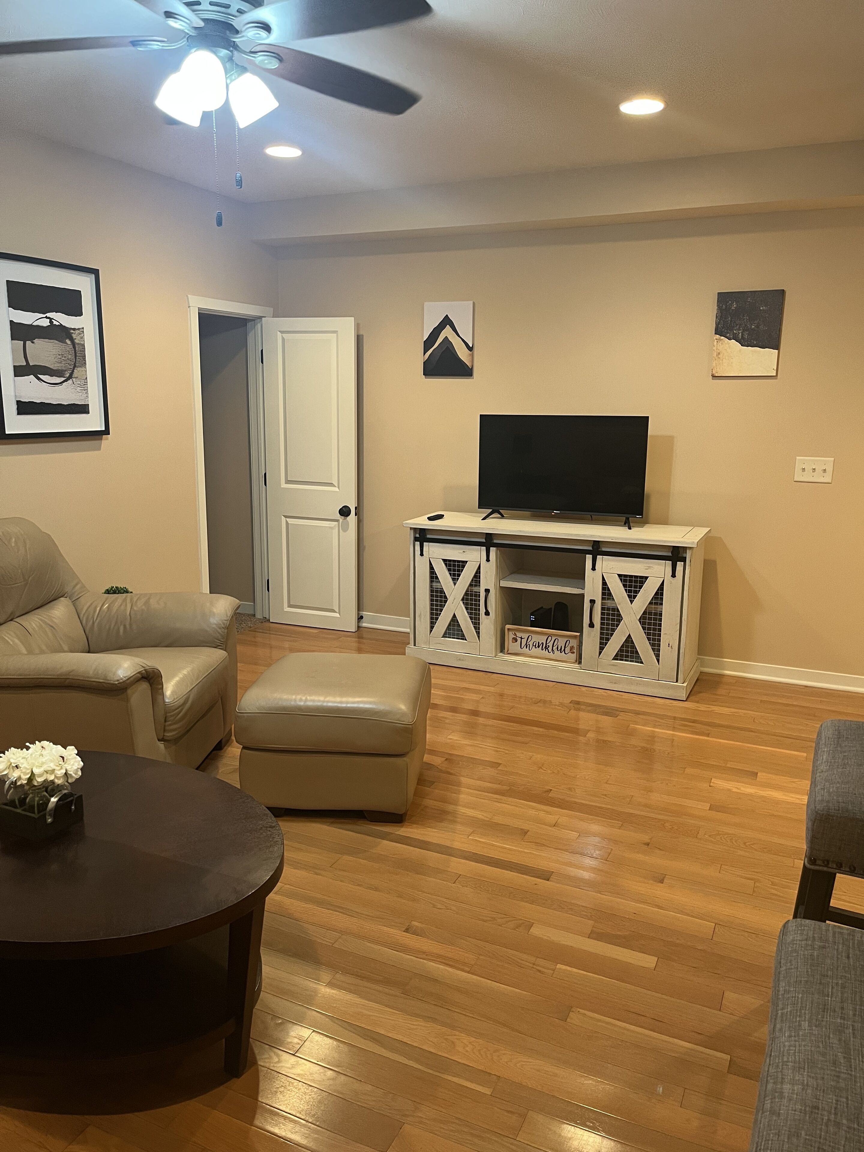 A living room with a beige sofa, a coffee table, a TV on a white entertainment unit, and a ceiling fan.