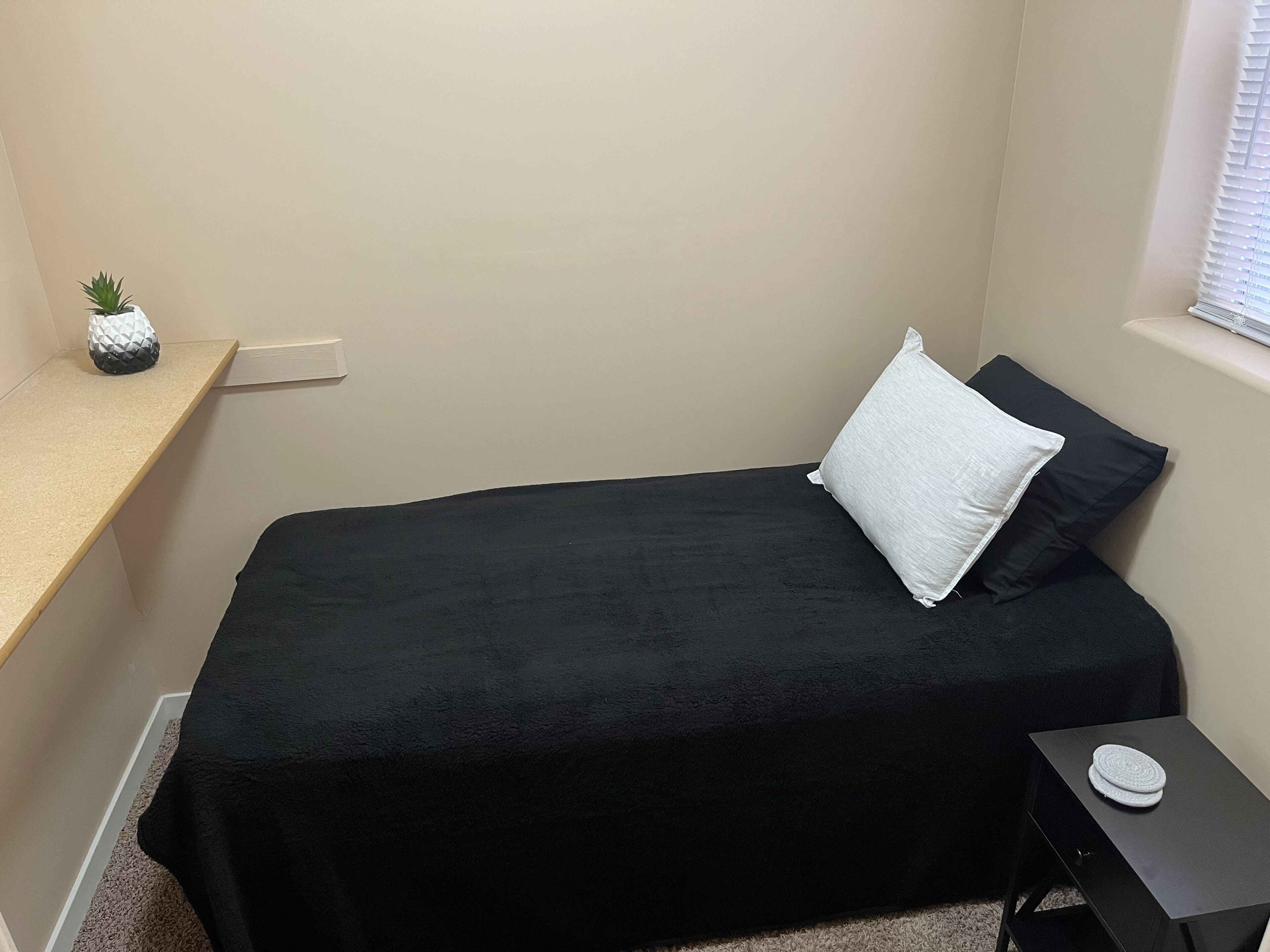 A small, neatly arranged bedroom with a single bed, a nightstand, a small table, and a window with blinds.