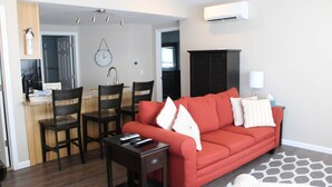 Ein Wohnzimmer mit rotem Sofa, zwei Sesseln, einem Couchtisch und einem Fernseher. Es gibt eine Küchenzeile mit Kühlschrank, Mikrowelle und einer Wanduhr.