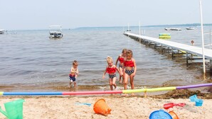 A beach with a pier extending into the water, a sandy shore with beach toys, and a few boats in the background. - Jack's on the Bay, 2 Bedroom/2 Bath Lake Front (Denton Township)