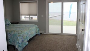 A bedroom with a bed, a window with blinds, a door, and a view of the beach. - Jack's on the Bay, 2 Bedroom/2 Bath Lake Front (Denton Township)
