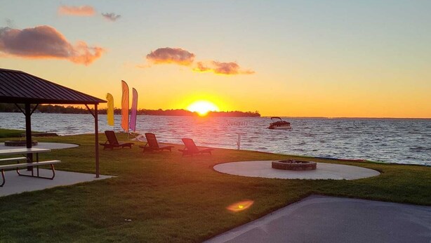 A sunset view from a lakeside patio with lounge chairs, a picnic table, and a small boat on the water. - Jack's on the Bay, 2 Bedroom/2 Bath Lake Front (Denton Township)