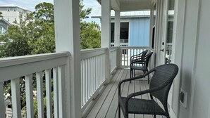 Eine überdachte Veranda mit weißen Geländern, zwei Korbstühlen und Blick auf ein blaues Gebäude im Hintergrund.