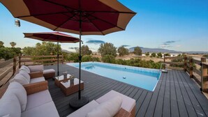 Zona junto a la piscina con terraza de madera, mobiliario de mimbre y un paraguas rojo.