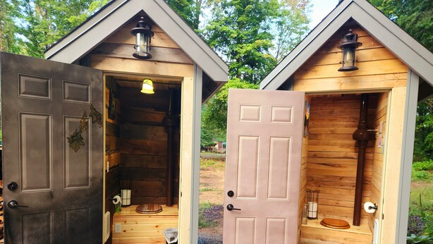 Deux cabanes en bois, dont les portes sont ouvertes, et qui sont chacune équipées de toilettes, d’un petit lavabo et d’une poubelle.