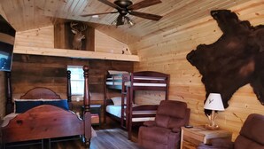 Une chambre rustique dans un chalet, avec un plafond en bois, un ventilateur de plafond, un lit superposé, une chaise en bois, une petite table avec une lampe et une peau d’animal accrochée au mur.