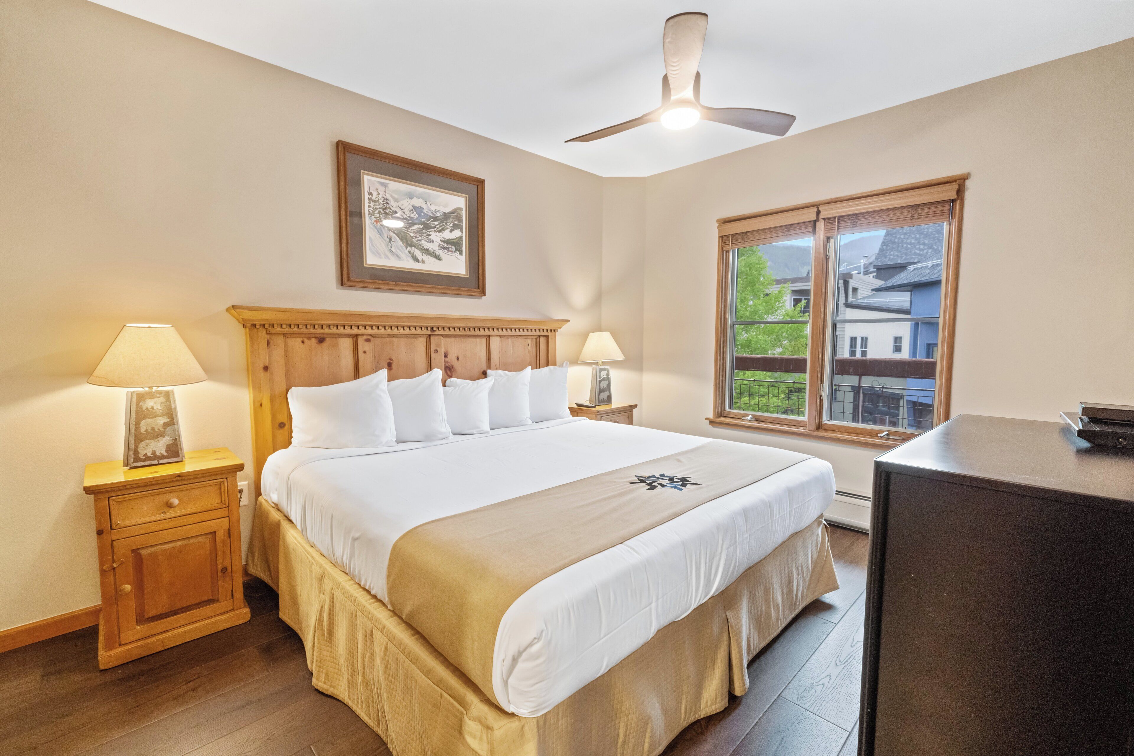 A bedroom with a large bed, wooden headboard, two bedside lamps, a ceiling fan, a window with a view, and a desk.