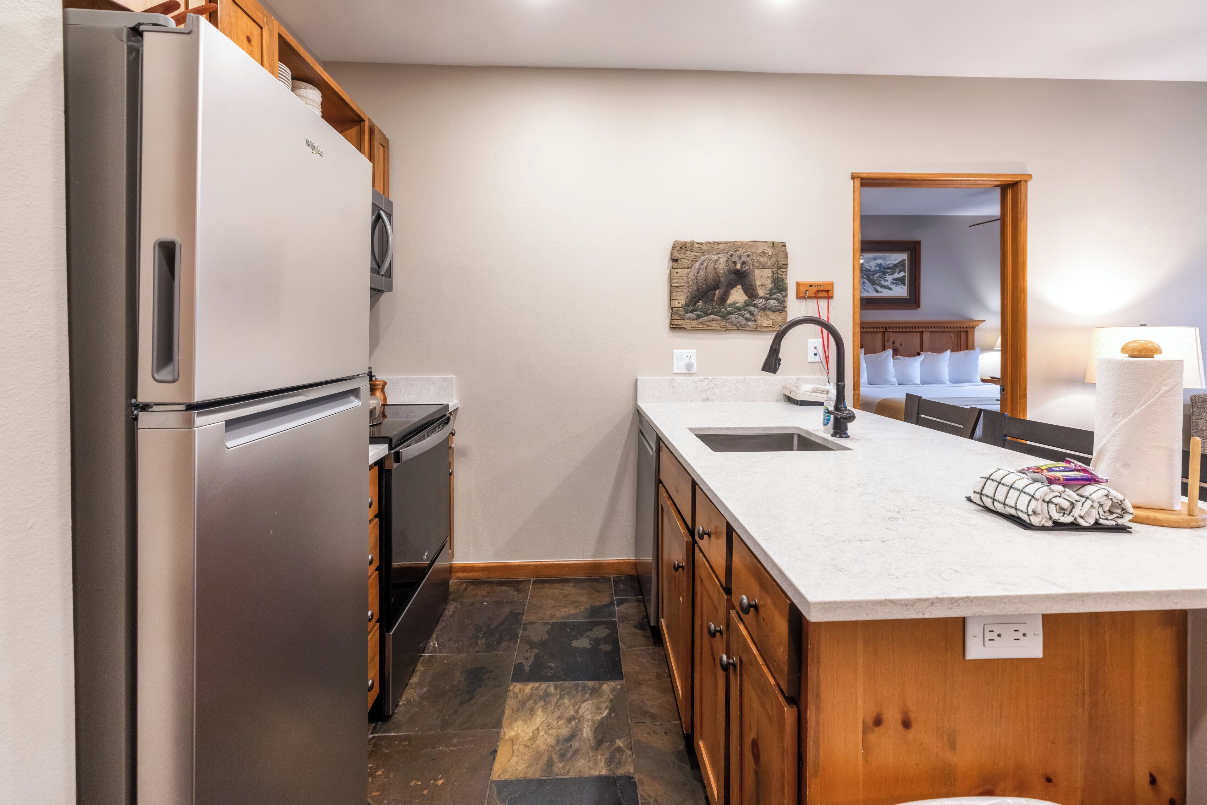 A modern kitchen with a stainless steel refrigerator, wooden cabinets, a marble countertop, and a sink.