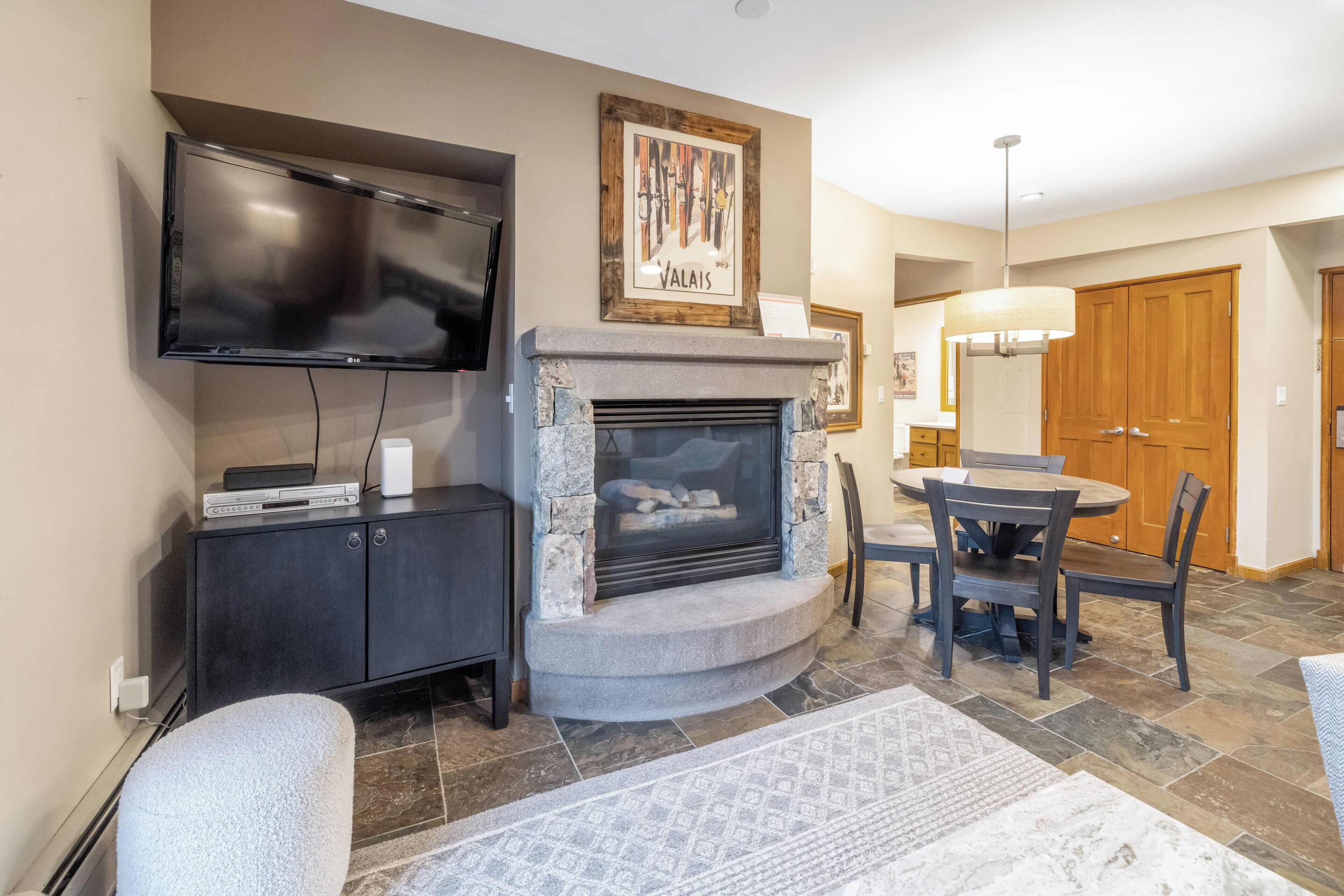 A living room with a fireplace, a flat-screen TV mounted on the wall, a dining table with chairs, and a patterned rug.