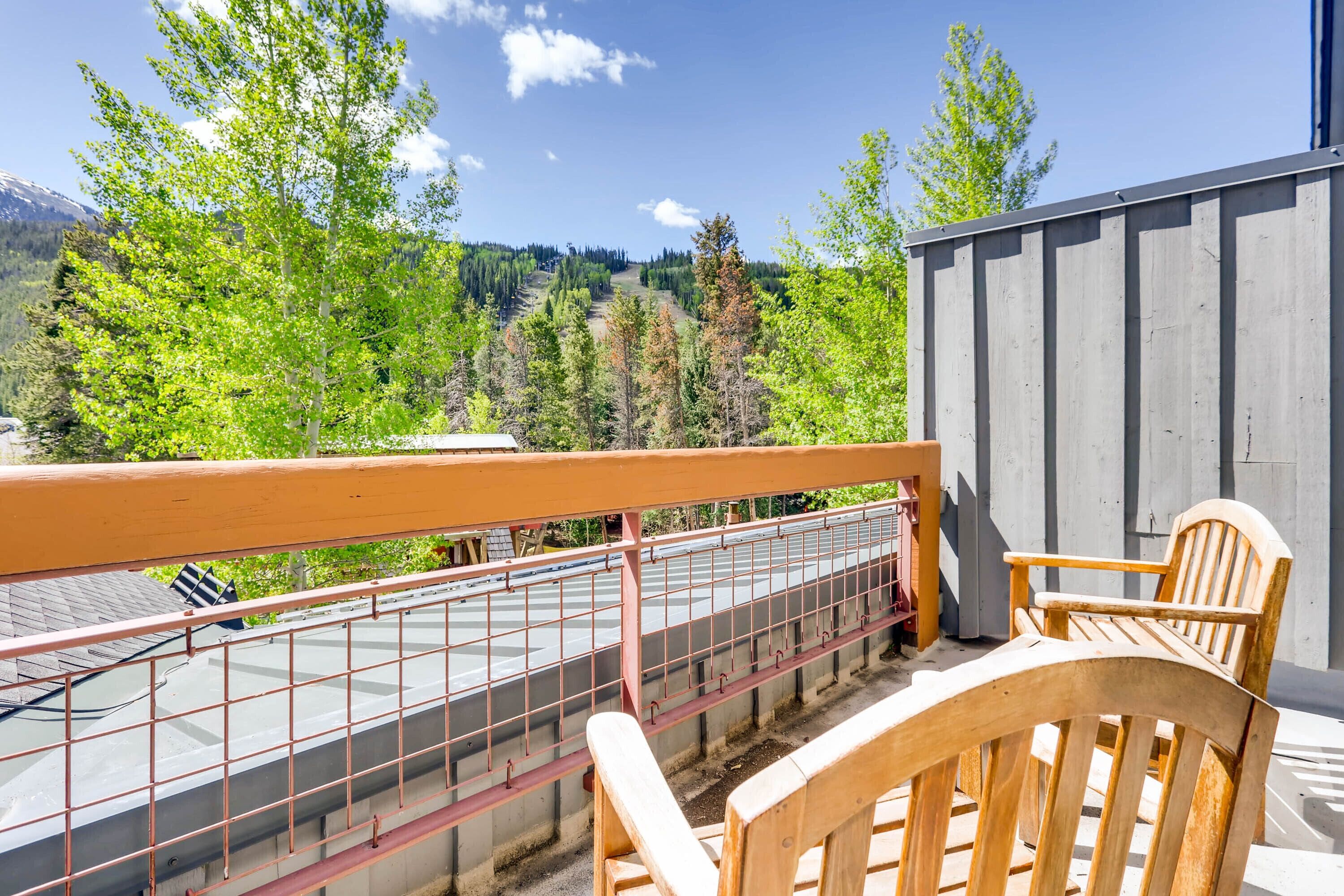 A balcony with a wooden chair and a metal railing overlooking a mountainous landscape with trees and a building in the background.
