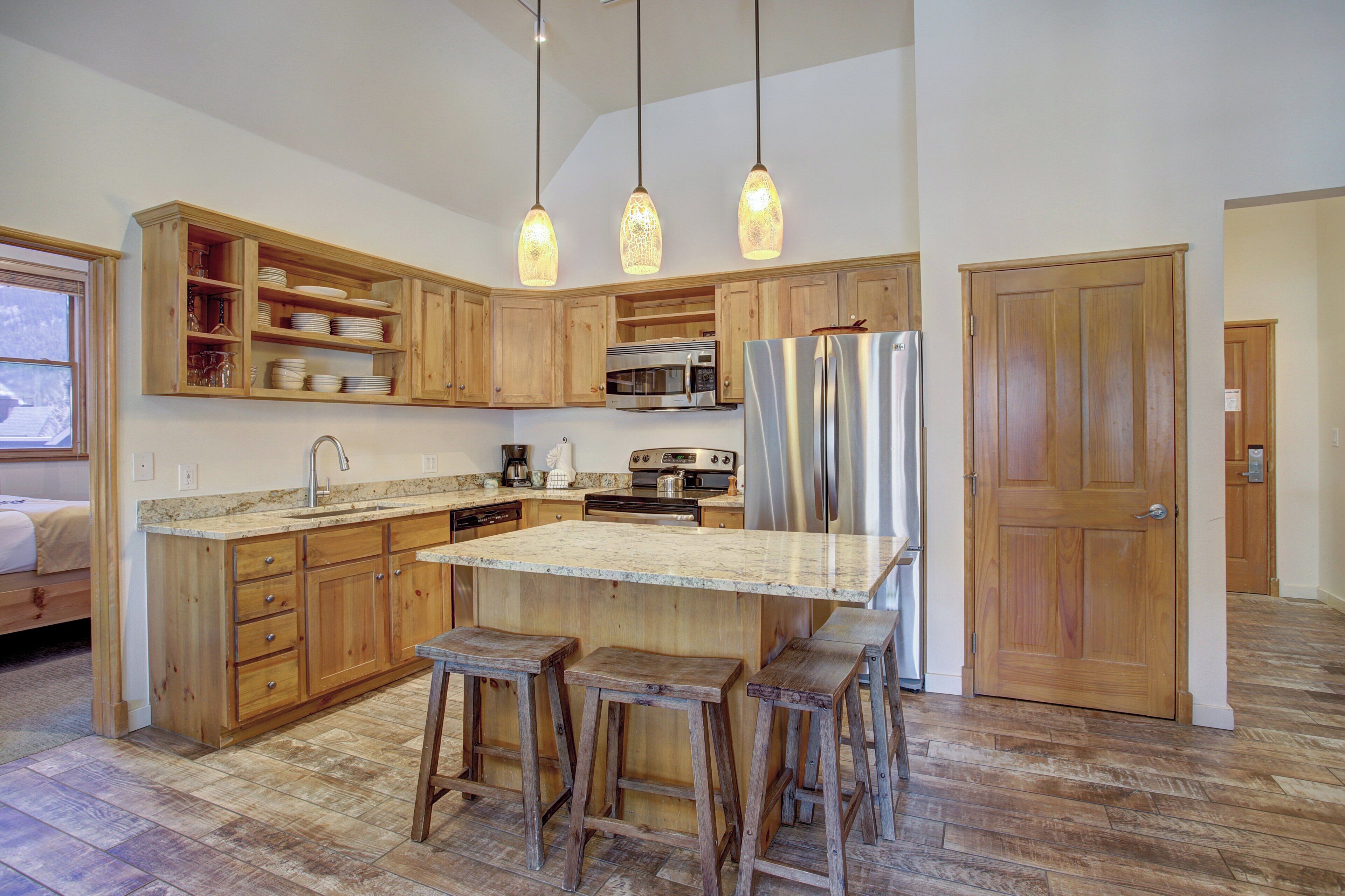 A kitchen with wooden cabinets, a large island with stools, a stainless steel refrigerator, and pendant lights.