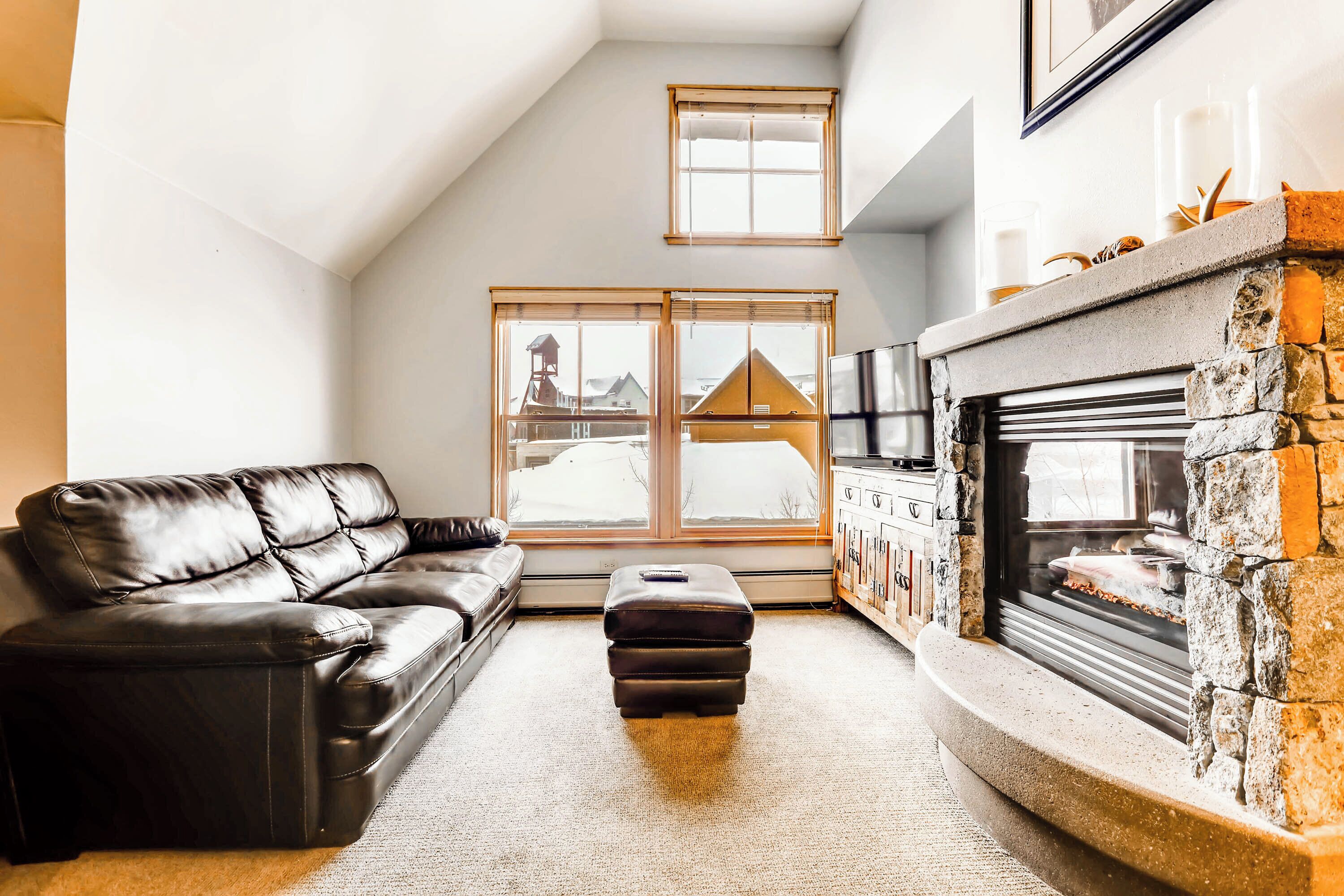 A cozy living room with a black leather sofa, a matching ottoman, a stone fireplace, and large windows offering a view of the outside.