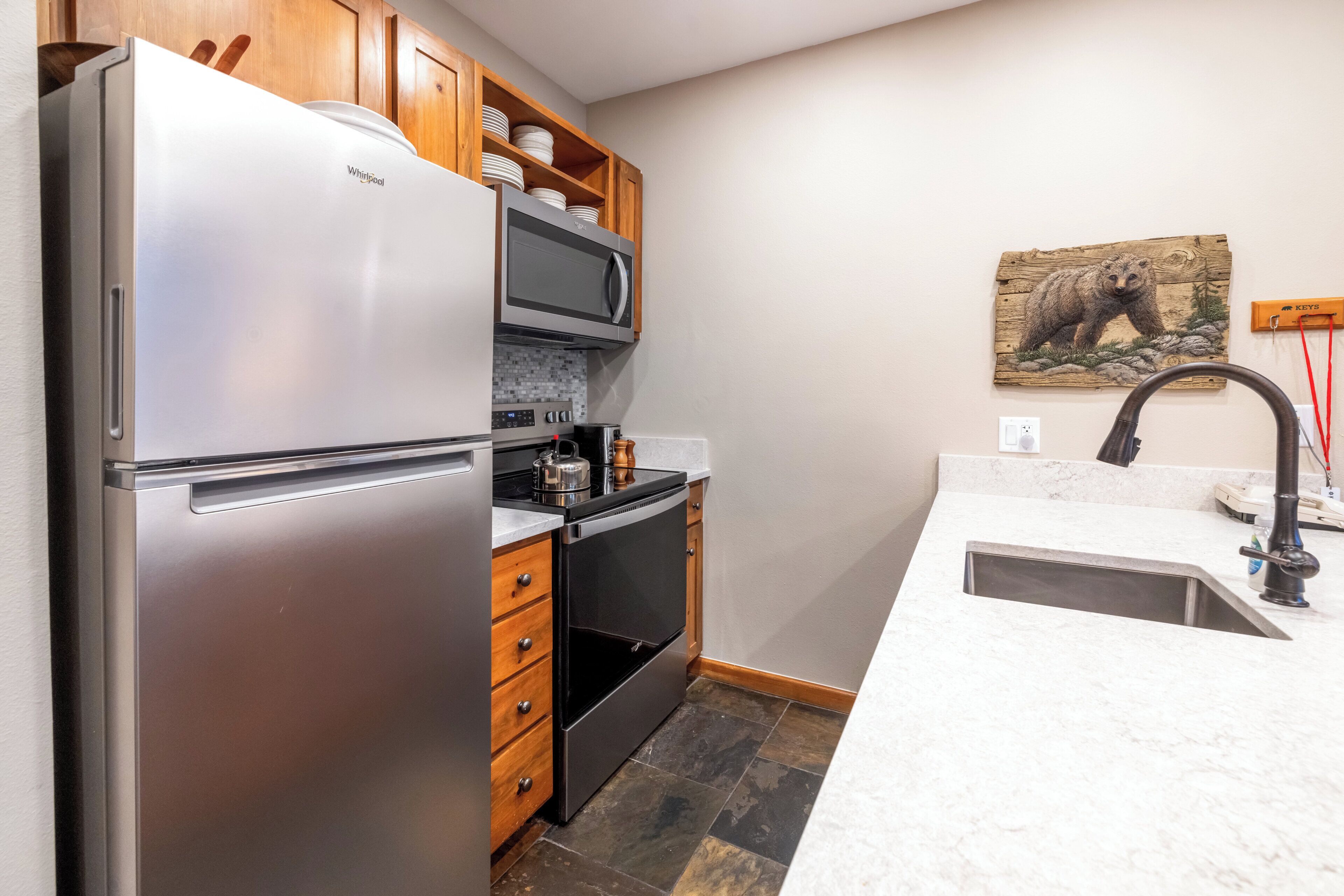A kitchen with a stainless steel refrigerator, a microwave, wooden cabinets, a tiled backsplash, and a bear-themed artwork on the wall.