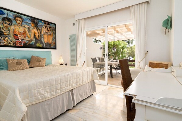 A bedroom with a bed, a painting, a dining area with a table and chairs, and a view of the outdoors.