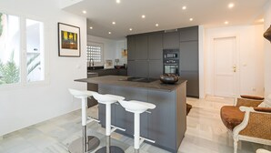 Una cocina moderna con una isla central, taburetes de bar y un comedor con sillas de mimbre.