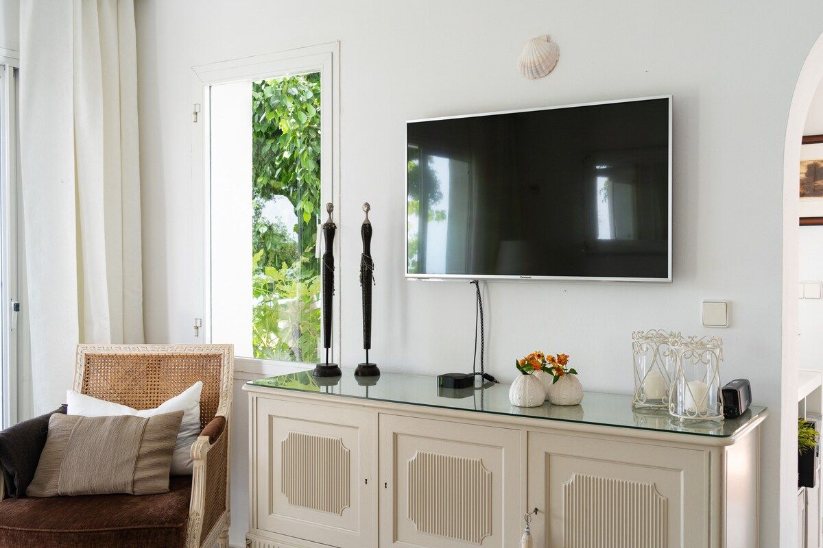 Una habitación con una televisión montada en la pared, un aparador de cristal, una silla de mimbre y una puerta que da a un jardín.