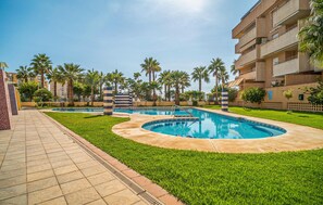 A swimming pool area with a tiled walkway, palm trees, and a multi-story building in the background. - Lovely apartment in Motril (Motril)