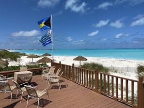 A wooden deck with lounge chairs and a table, overlooking a beach with clear turquoise water and a flagpole with flags. - Beach Villa 675 Steps to the pool and beach, 2 king bedrooms with private baths (Treasure Cay)