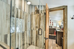 Une douche moderne avec un revêtement mural en bois et une porte en verre, donnant sur un espace cuisine équipé d’un évier et d’un plan de travail.