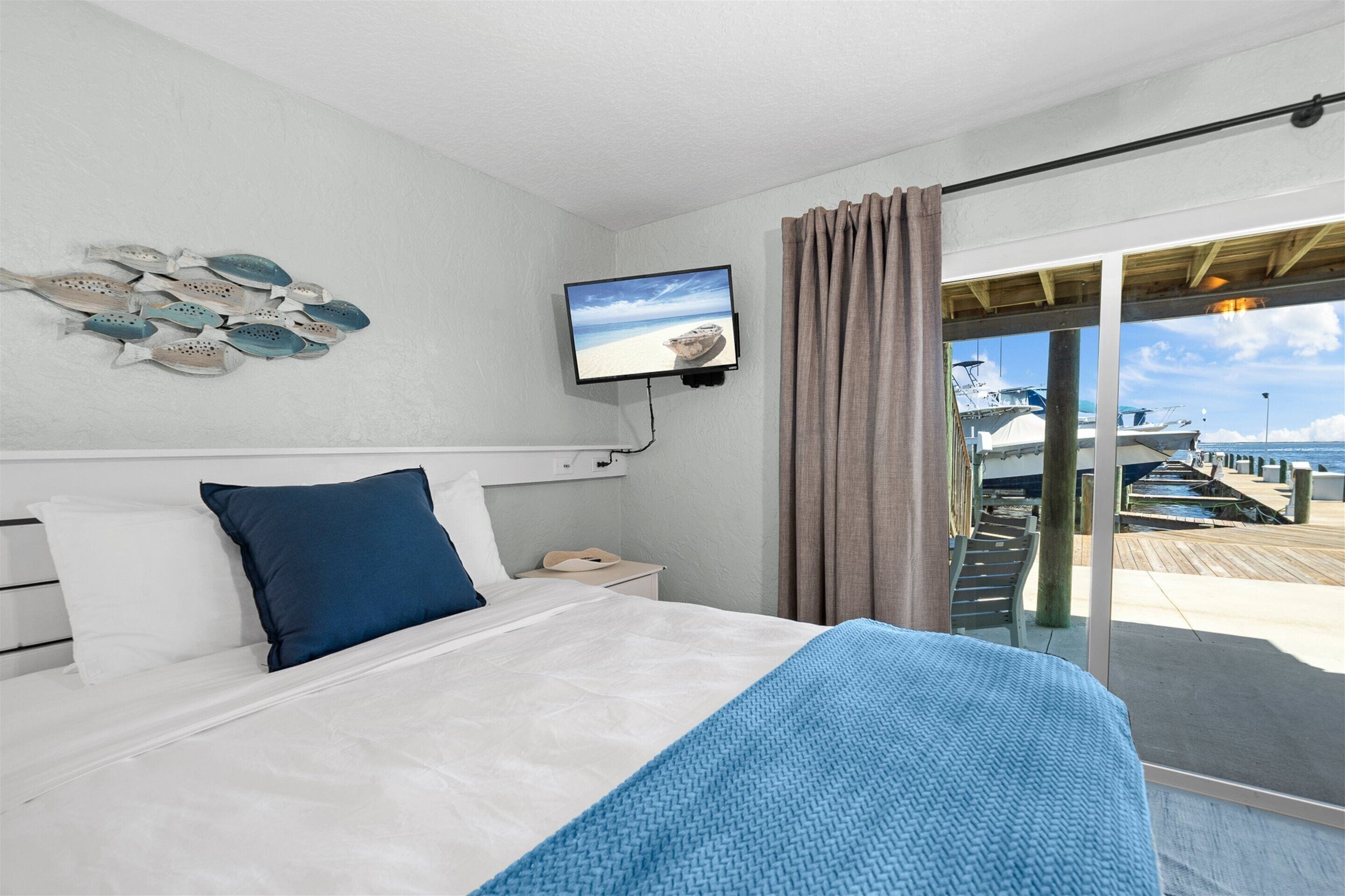 A bedroom with a bed, a TV mounted on the wall, a nightstand, and a view of a dock and boats through a sliding glass door.