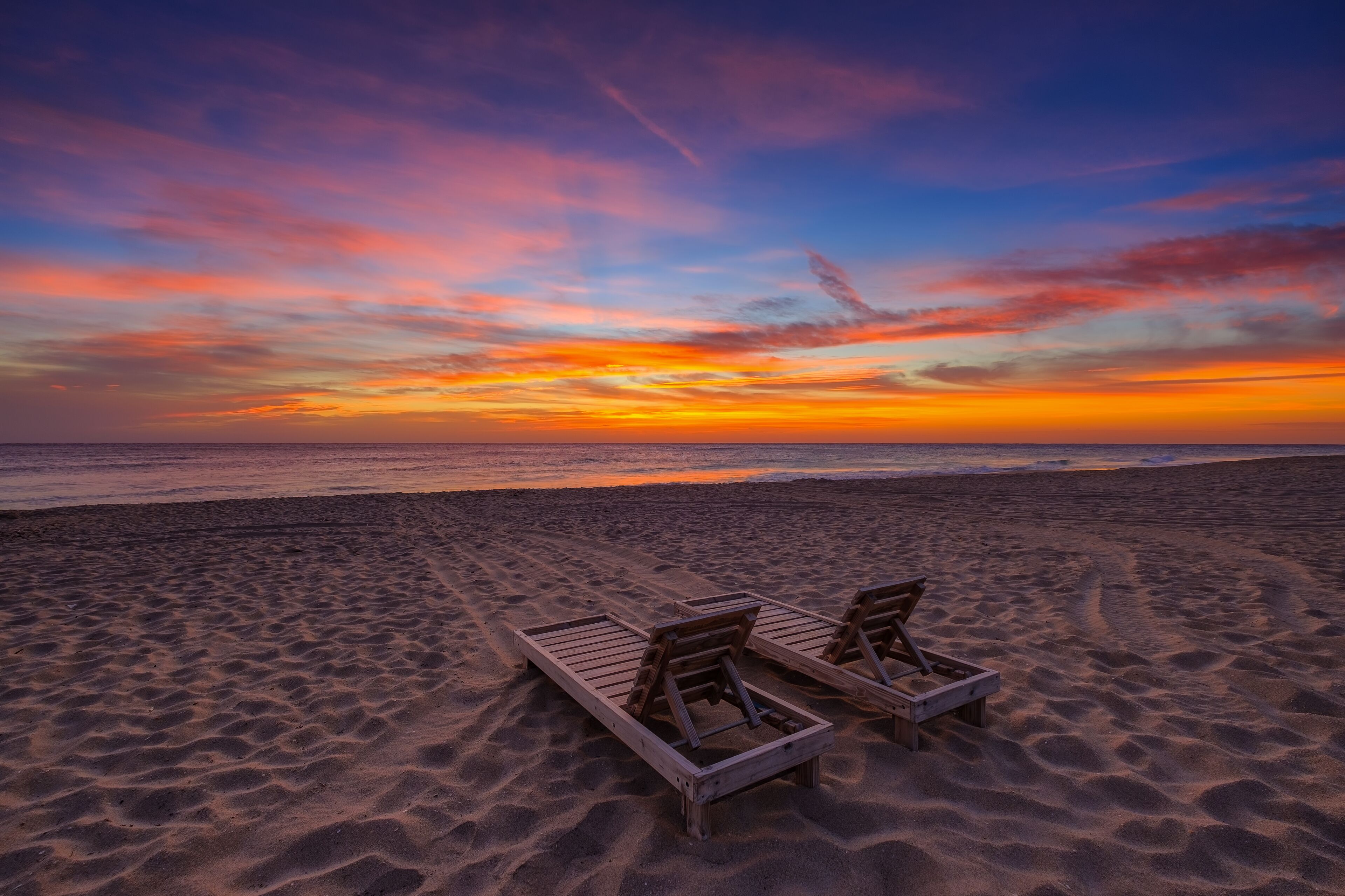 A beach with two wooden lounge chairs facing the ocean during sunset.
