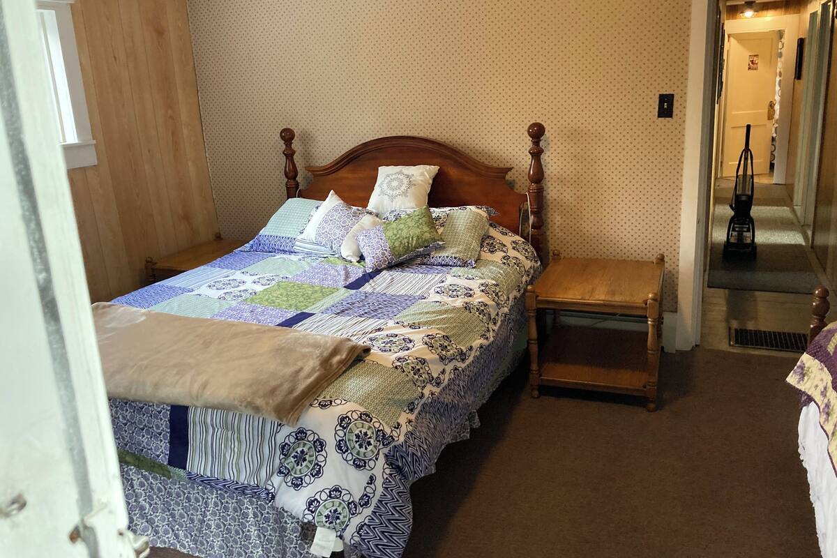 Una cama bien hecha con un edredón estampado, una mesita de noche de madera y una puerta que da a un pasillo.