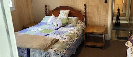 Una cama bien hecha con un edredón estampado, una mesita de noche de madera y una puerta que da a un pasillo.