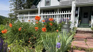 A white house with a porch, surrounded by a garden with various flowers and plants. - Seymour Lake Lodge (Morgan)